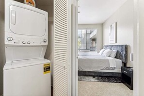 Een wasruimte met een voorlader wasmachine en droger, een bed met een grijze hoofdbord, en een raam met jaloezieën.