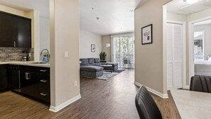 A modern kitchen with dark cabinetry and a tiled backsplash, a dining area with a table and chairs, and a living room with a gray sectional sofa and a large window. - Cozy Marina Hideaway Gym, Pool & Spa Central (Marina del Rey)