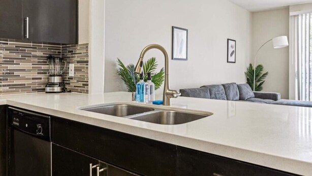 A modern kitchen with a double sink, a built-in dishwasher, and a coffee machine. There is a grey sofa and a floor lamp in the living area. - Cozy Marina Hideaway Gym, Pool & Spa Central (Marina del Rey)