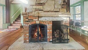A stone fireplace with a fire, a wooden staircase, and a view through the windows. - Old Orchard House (Brandon)