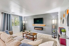 A modern living room with a sofa, coffee table, fireplace, TV, and bookshelves. - Comfortable 2BR Apt Near Capitol Hill & SLC (Salt Lake City)