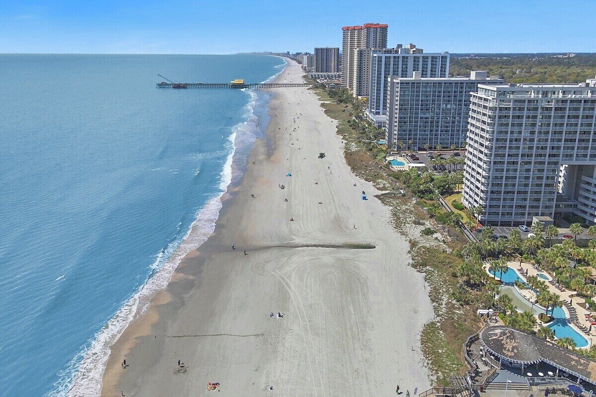 A beach with a pier extending into the ocean, surrounded by high-rise buildings and a resort area with pools.