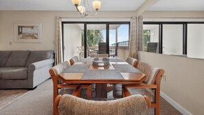 A spacious living room with a large dining table, comfortable seating, a chandelier, and a view through large windows. - Ocean Bridge 1A2 (Myrtle Beach)