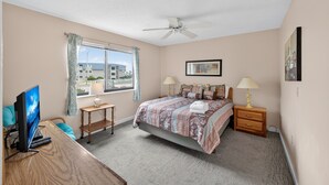 A bedroom with a bed, bedside tables, a desk with a computer, a ceiling fan, and a window with curtains. - Ocean Bridge 1A2 (Myrtle Beach)