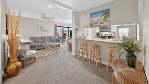 A modern living room with a grey sofa, a coffee table, and a dining area with stools. - Ocean Bridge 1A2 (Myrtle Beach)