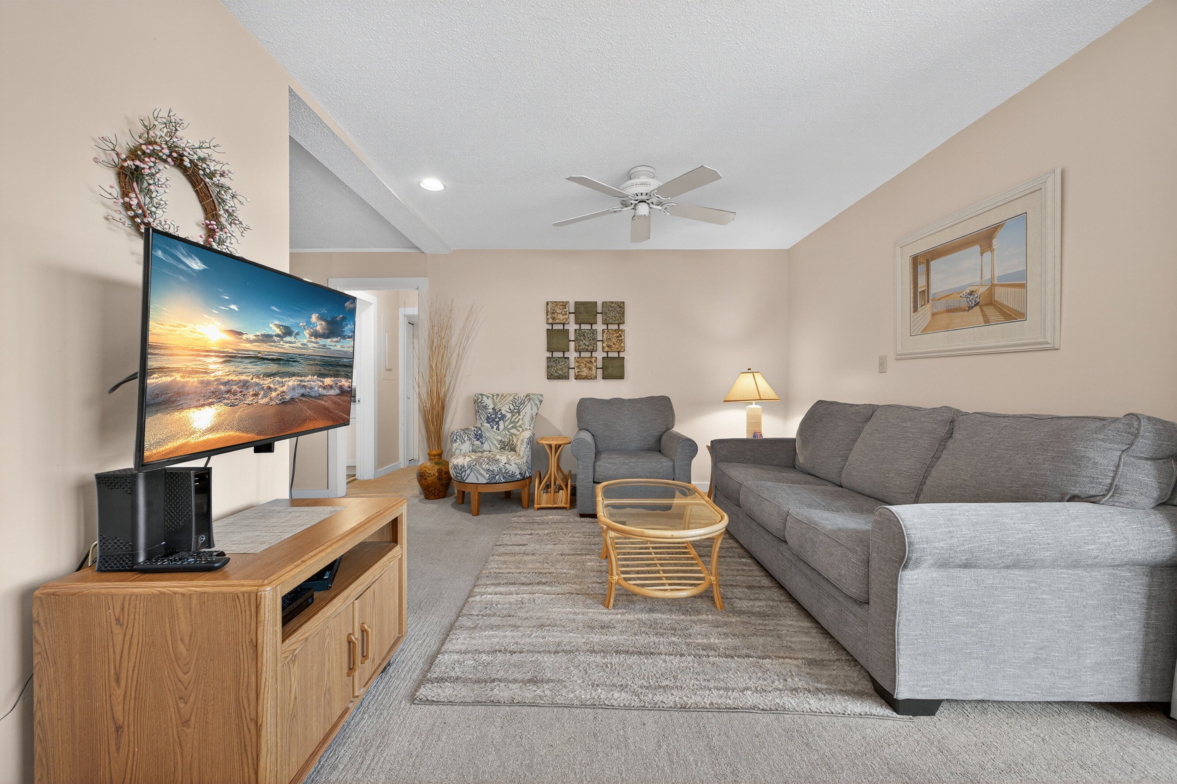 A living room with a grey sofa, a television on a wooden stand, a ceiling fan, and a framed picture on the wall.