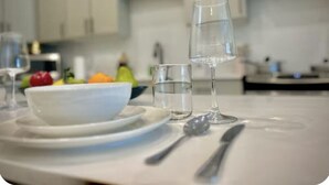 A dining table with a bowl of fruit, a glass of water, and a wine glass in a modern kitchen with white cabinets and a stainless steel backsplash. - Workerfriendly, Washerdryer, Wifi,longstaydiscount (Sherman)