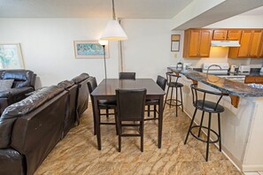 A modern kitchen with wooden cabinets, a dining area with a table and chairs, and a bar area with stools. - Sands Beach Club 120 (Myrtle Beach)