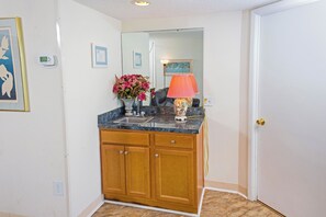 A bathroom with wooden cabinets, a marble countertop, a sink, a mirror, a vase with flowers, and a lamp. - Sands Beach Club 120 (Myrtle Beach)