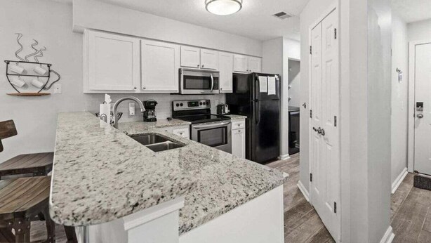 A modern kitchen with white cabinets, a granite countertop, stainless steel appliances, and a wooden dining table. - Maison Place I (Myrtle Beach)