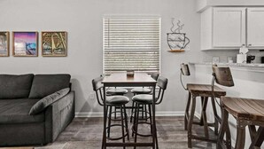 A modern living room with a gray sofa, a wooden dining table with four stools, and a kitchen area with white cabinets and a hanging light fixture. - Maison Place I (Myrtle Beach)