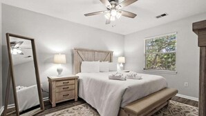 A bedroom with a bed, bedside tables, a ceiling fan, a large mirror, and a window with blinds. - Maison Place I (Myrtle Beach)
