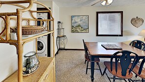A room with a dining table, chairs, a washing machine, a shelf with baskets, a decorative wall piece, and a ceiling fan. - Shipwatch Pointe I F217 (Myrtle Beach)