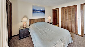 A bedroom with a bed, two bedside tables with lamps, a nightstand, a wooden headboard, a painting on the wall, and a door with a wooden shutter. - Shipwatch Pointe I F217 (Myrtle Beach)