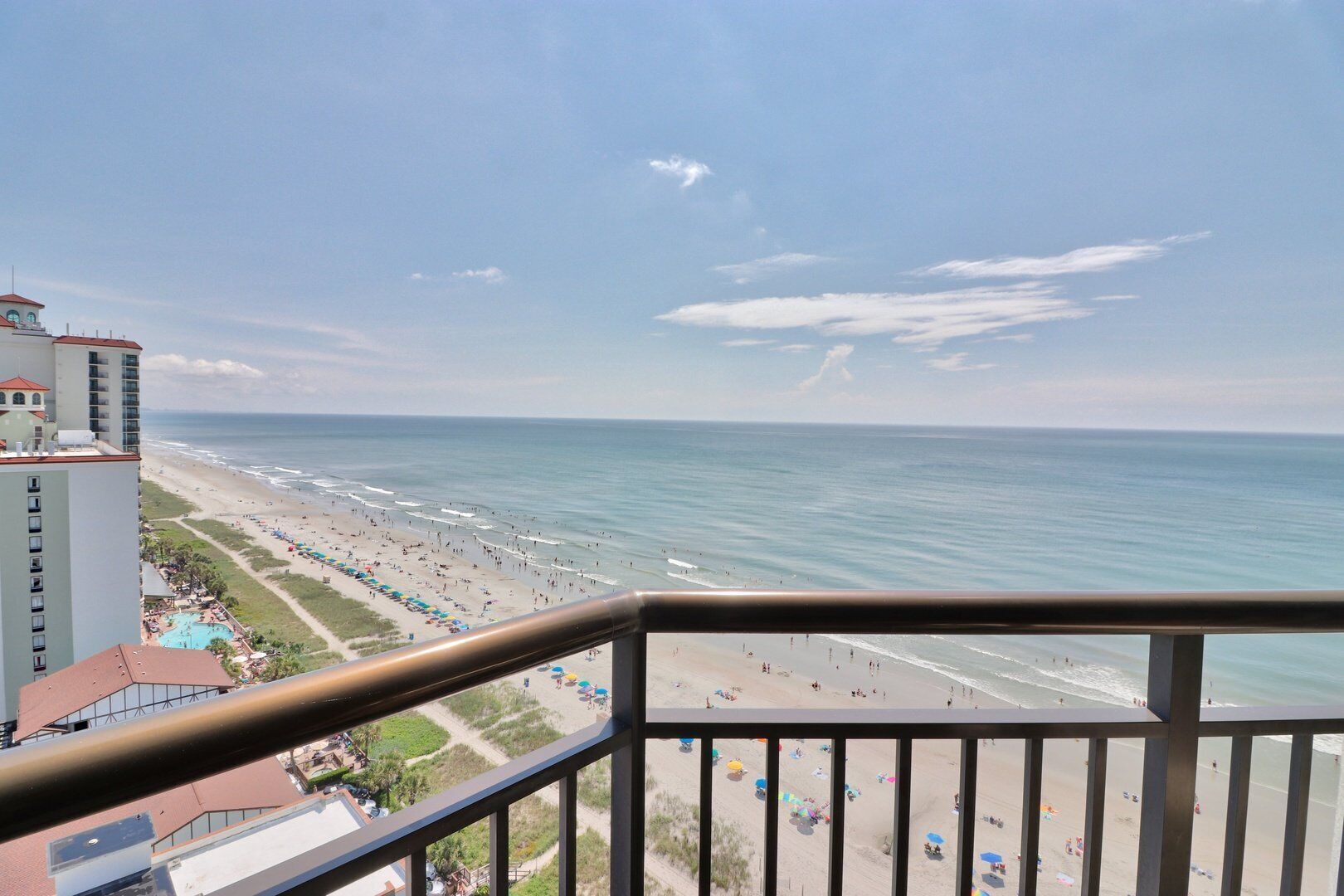 A beach view from a balcony with a railing, a pool, and beach umbrellas.