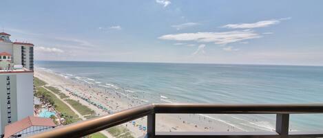 A beach view from a balcony with a railing, a pool, and beach umbrellas.