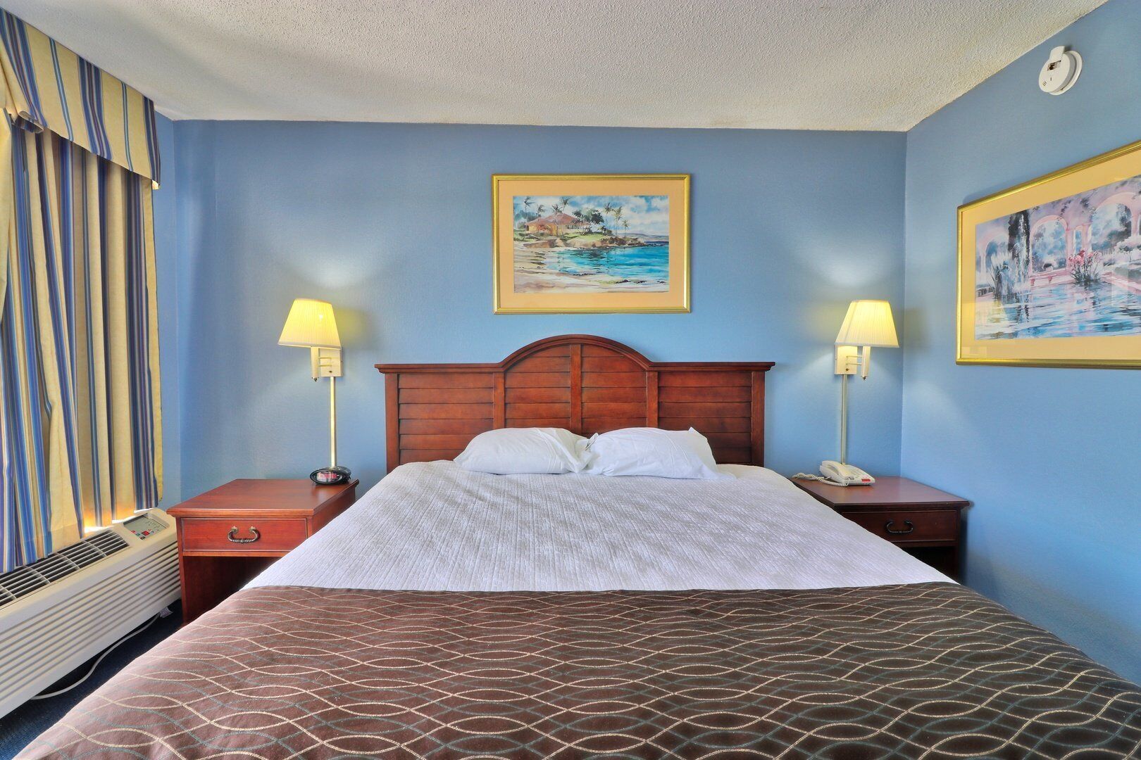 A hotel room with a bed, two bedside lamps, a heater, and two framed pictures on the walls.