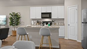 A modern kitchen with white cabinets, a microwave, and a sink. There are two bar stools and a dining table with chairs. A potted plant and a television are also visible. - Marbella Lane - Amazing 4BR Lakeview Home (Austin)