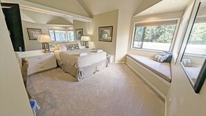 Ein Schlafzimmer mit einem großen Bett, einem Nachttisch, einer Kommode, einer Sitzgelegenheit am Fenster und Blick ins Freie.