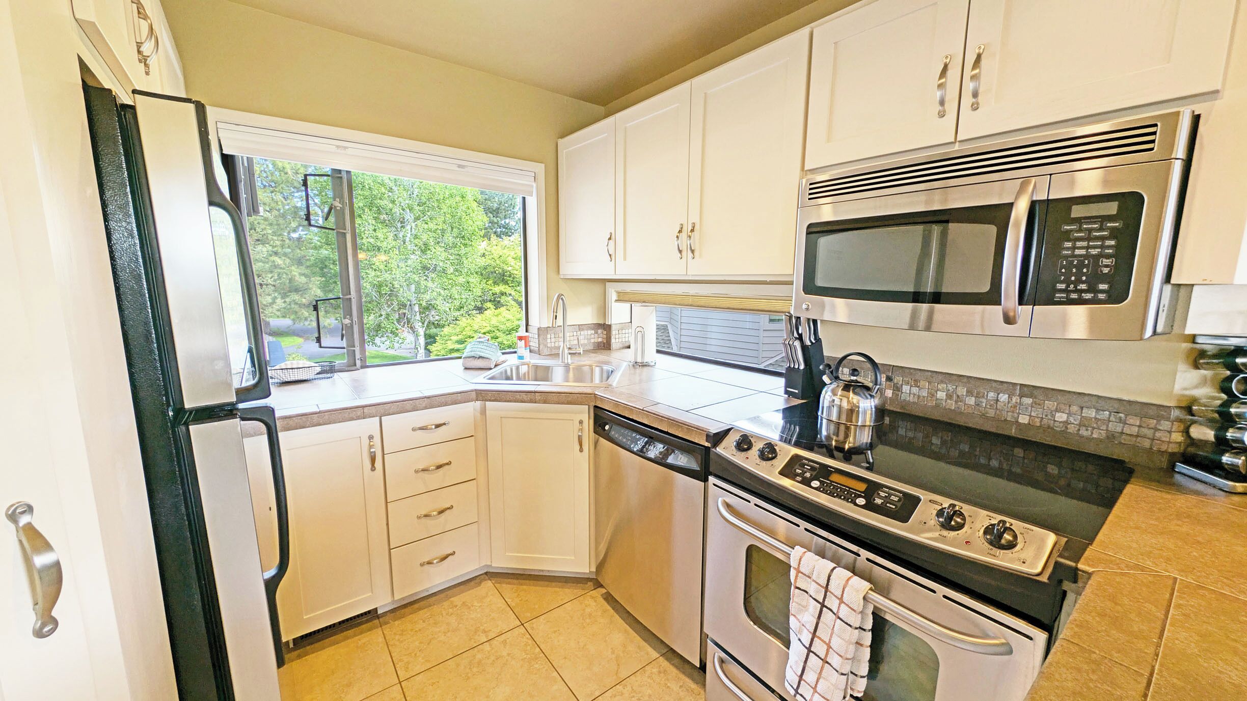 A modern kitchen with white cabinets, stainless steel appliances, and a window providing a view of greenery.
