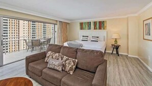 A hotel room with a bed, a brown sofa, a small table, a lamp, and a balcony with a view of high-rise buildings. - Marbella Lane - Ocean Marina View Ilikai Hotel (Honolulu)