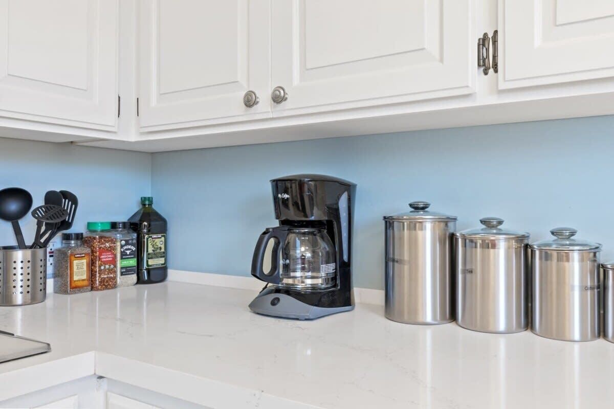 Una cocina con gabinetes blancos, cafetera, frascos de especias y recipientes de acero inoxidable.