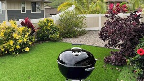 Ein Garten mit einem schwarzen Grill, grünem Rasen und einem Haus im Hintergrund.