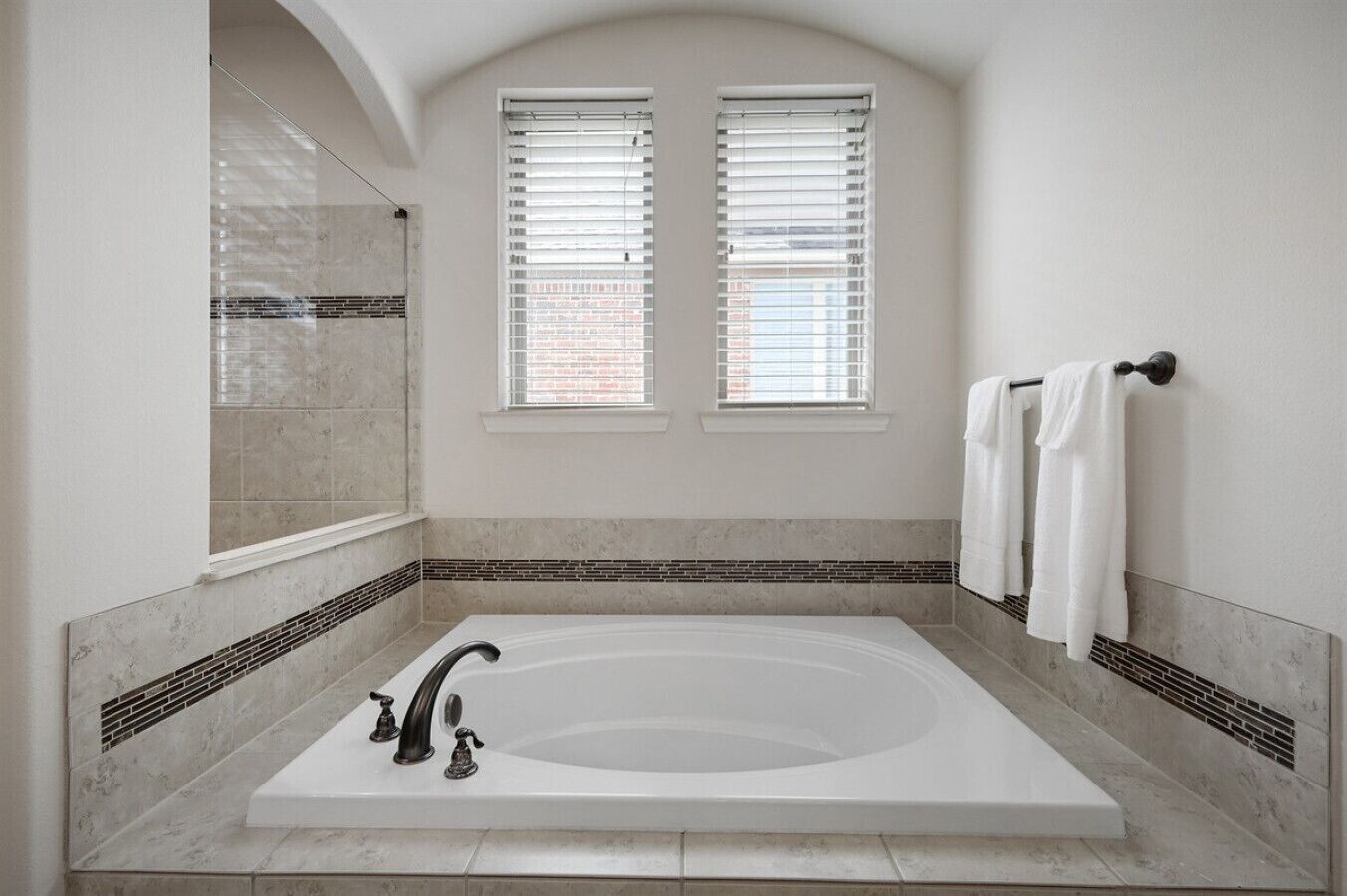 A large, rectangular bathtub with a single faucet, flanked by two windows with blinds, in a room with a tiled floor and walls.