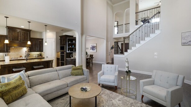 Een ruime woonkamer met een grote bank, twee fauteuils en een salontafel. In de achtergrond is een keuken zichtbaar met donkerhouten kastjes en hanglampen.