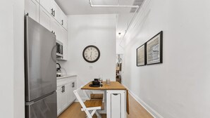 A narrow kitchen with white cabinetry, a stainless steel refrigerator, a wooden dining table with a chair, a wall clock, and framed artwork. - 2BR 2BA in Historic Brownstone Near Subway (New York)
