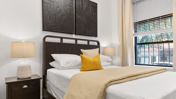 A neatly made bed with white linens and a mustard-colored throw, a nightstand with a lamp, and a window with blinds. - 2BR 2BA in Historic Brownstone Near Subway (New York)