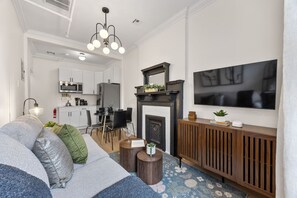 Uma sala de estar moderna com sofá, mesa de centro e lareira. Há uma área de jantar com cadeiras e uma cozinha ao fundo.
