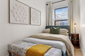 Quarto com uma cama, um banquinho, uma mesinha de cabeceira com abajur e duas obras de arte penduradas na parede.