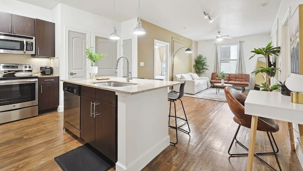 A modern kitchen with stainless steel appliances, a central island with a sink, and a dining area with a white table and brown chairs. - Luxurious 2b2b Near Toyota HQ, The Star, Legacy West (Frisco)