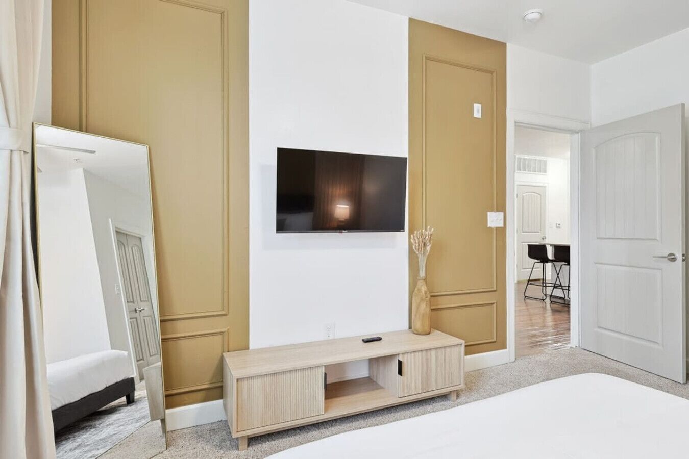 A modern bedroom with a bed, a TV mounted on the wall, a wooden cabinet, and a door leading to another room.