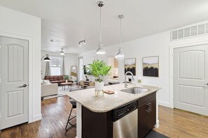 Cozinha moderna com bancada de granito, pia de aço inoxidável e lava-louças. Há uma área de refeições com sofá de couro marrom e mesa de centro.