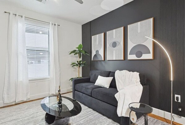 A modern living room with a dark grey sofa, a round glass coffee table, and a floor lamp. There are three framed artworks on the wall and a potted plant in the corner. - Chic and Stylish 2B Near Legacy West Wi-fi (Frisco)