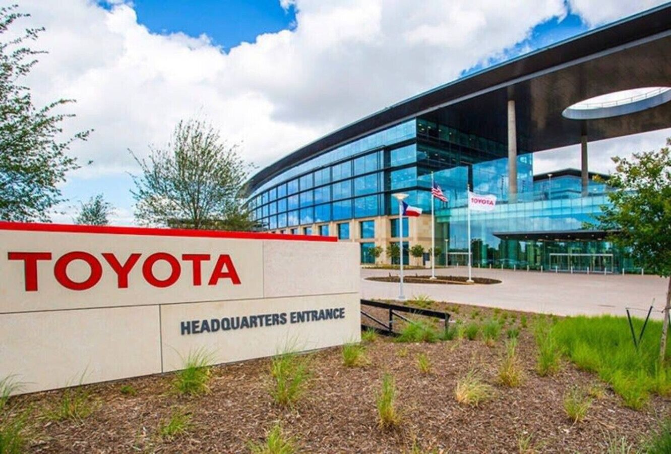 A Toyota corporate building with a modern glass facade, a sign indicating the headquarters entrance, and a landscaped area with grass and shrubs.