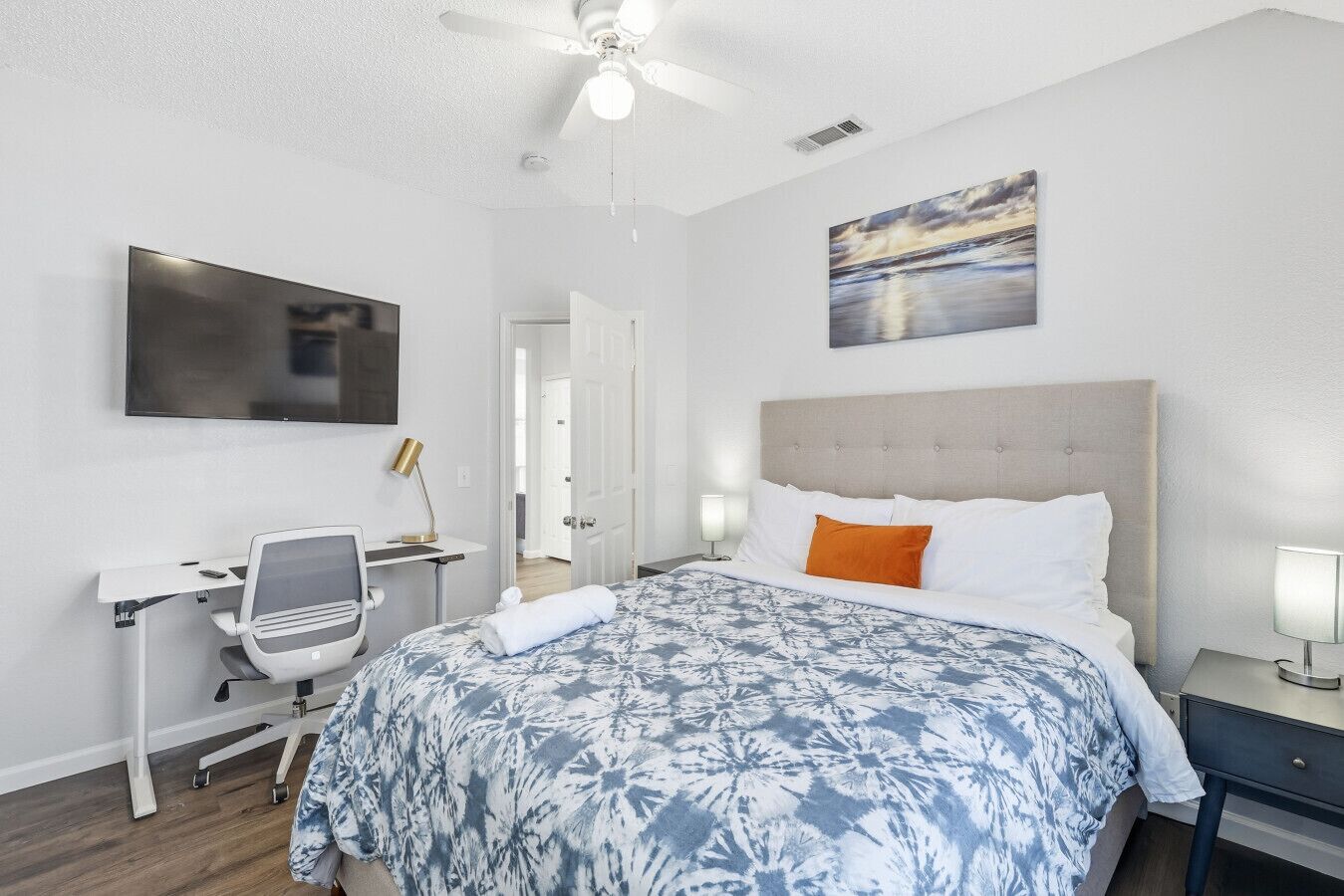 A hotel room with a bed, desk, chair, television, ceiling fan, artwork, and a door.