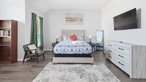 A modern bedroom with a bed, a desk, a dresser, a bookshelf, and a TV. - Bright & Cozy 4BR Home w Movie Room & Backyard (Frisco)