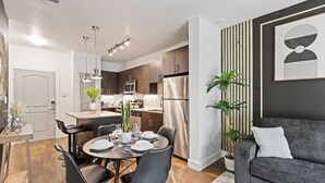 A modern kitchen with stainless steel appliances, a dining area with a round table and chairs, and a grey sofa in a living area. - Lux 1B w Pool Near Toyota HQ, The Star, Legacy West (Frisco)