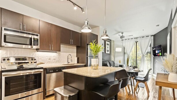 A modern kitchen with stainless steel appliances, a central island with bar stools, and a dining area with a view of the outdoors. - Lux 1B w Pool Near Toyota HQ, The Star, Legacy West (Frisco)