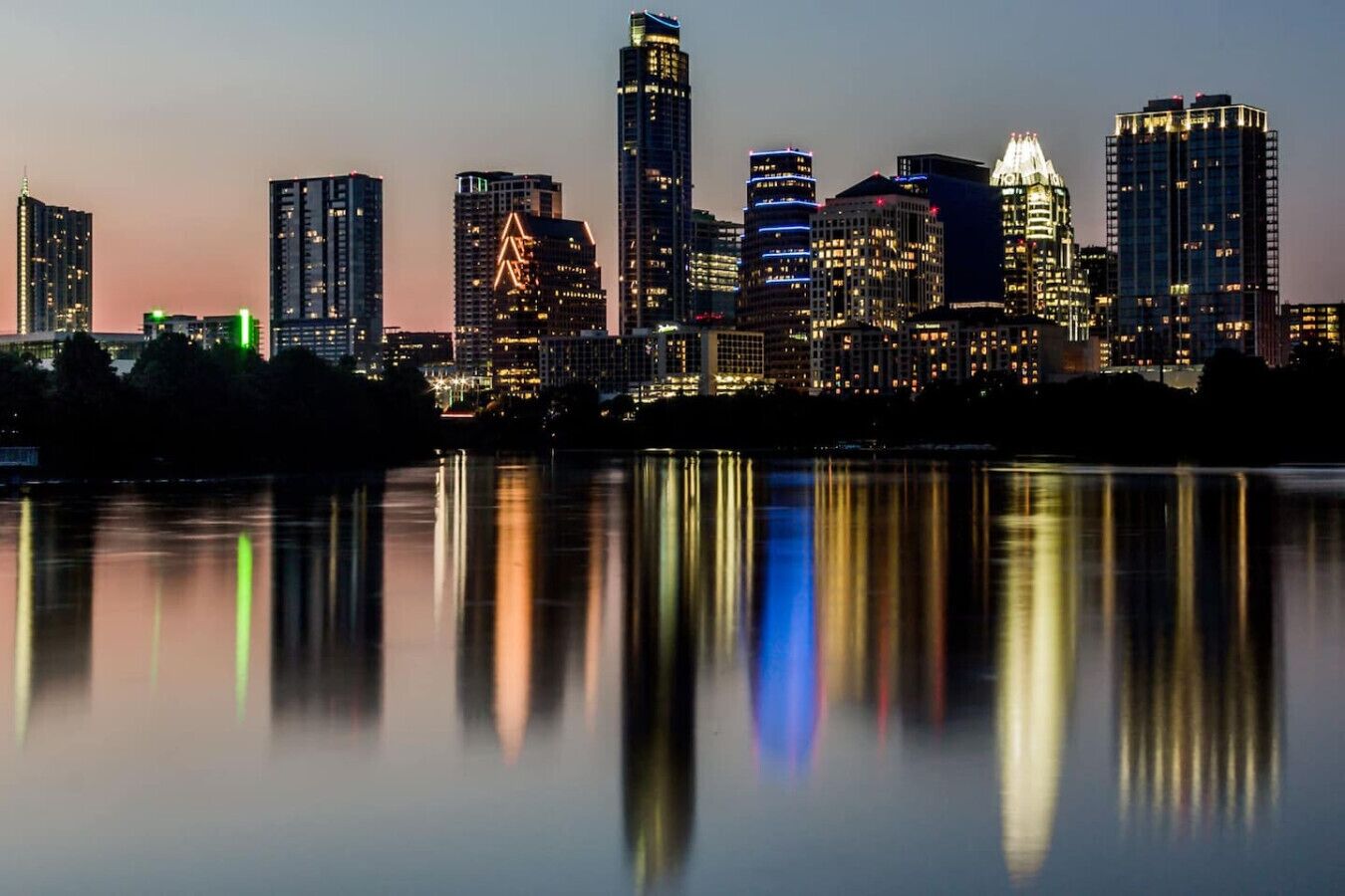 Un horizonte urbano al atardecer, con edificios iluminados reflejados en aguas tranquilas.