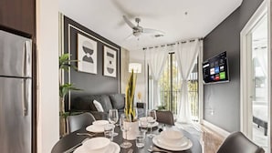 A modern dining area with a round table set for four, a ceiling fan, and a TV mounted on the wall. - Lux 1B w Pool Near Toyota HQ, The Star, Legacy West (Frisco)