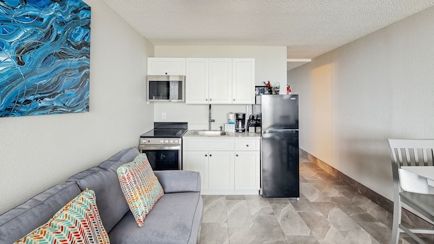 A compact living space with a kitchenette, grey sofa, patterned cushions, and a blue abstract painting on the wall.