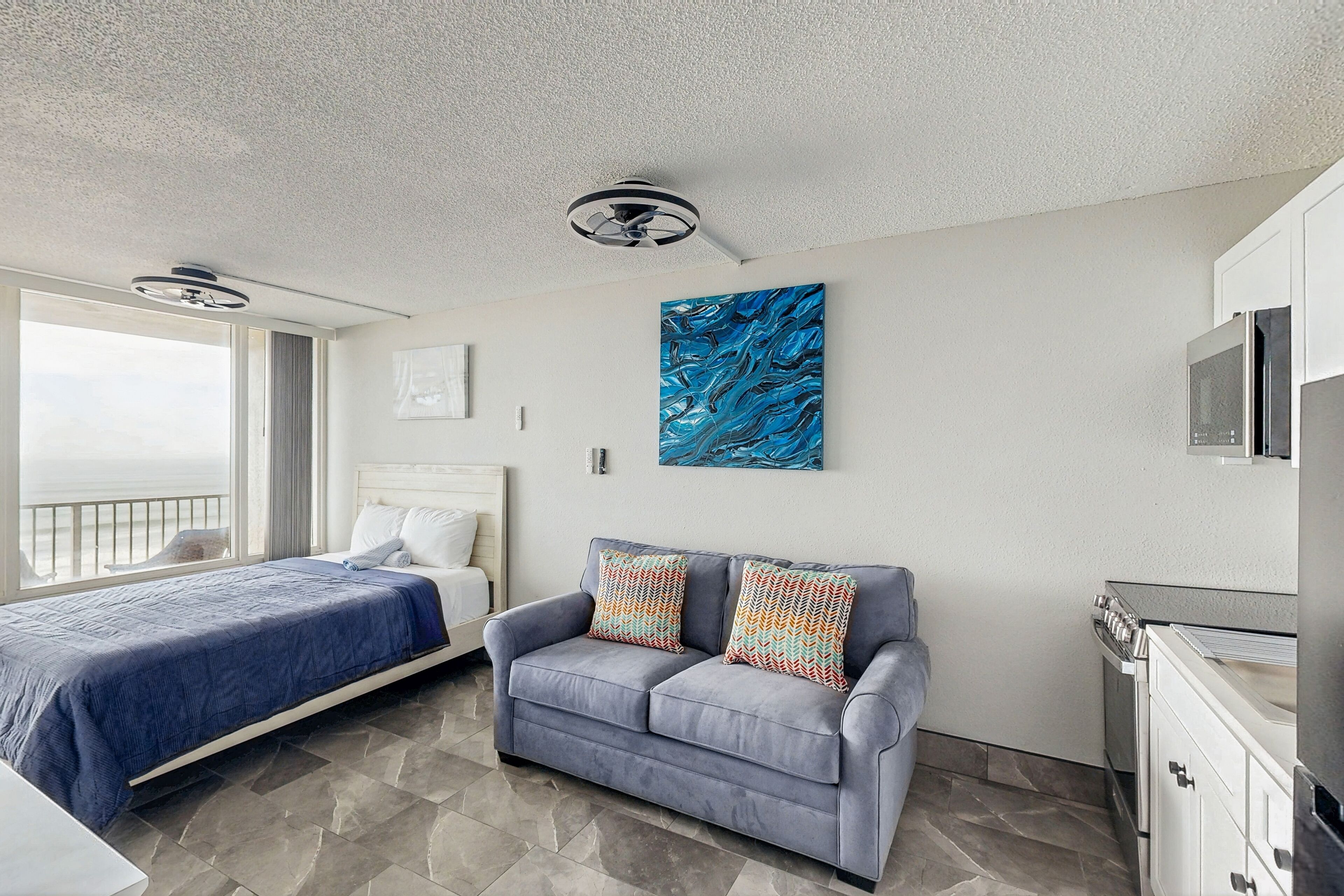 A modern bedroom with a bed, a gray sofa with patterned cushions, a ceiling fan, a large abstract painting, and a kitchenette.