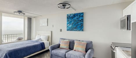 A modern bedroom with a bed, a gray sofa with patterned cushions, a ceiling fan, a large abstract painting, and a kitchenette.