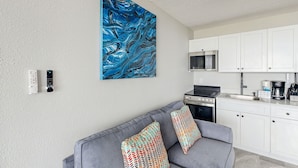A modern living room with a grey sofa, colorful cushions, a large abstract painting, a kitchen with white cabinets, and a built-in oven.