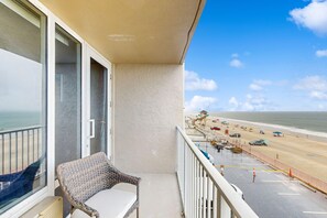 A balcony with a wicker chair, a view of the beach, and a parking area with beachgoers. - Pirates Cove 327 - Salt Air Cove (Daytona Beach Shores)