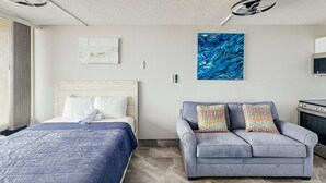 A hotel room with a bed, a gray sofa with patterned cushions, a blue abstract painting, a ceiling fan, and a microwave.